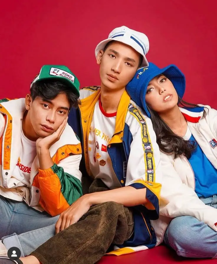 three models sitting closely together wearing colourful streetwear jackets and hats against a red background